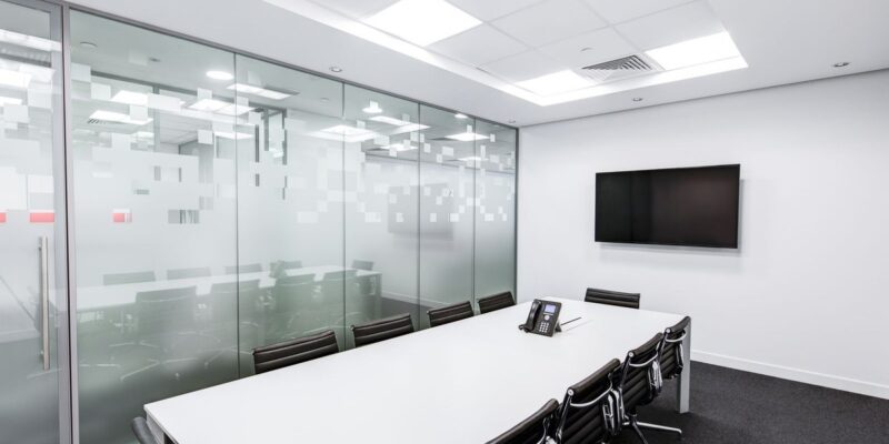 Empty modern conference room with a white table, black chairs, wall TV, and frosted glass partition