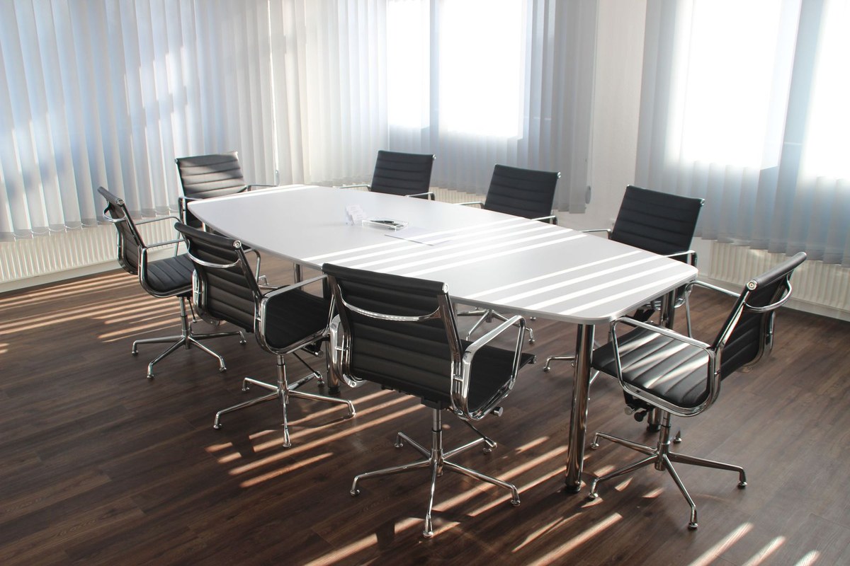 Sunlit conference room with a white boat-shaped table, eight black chairs, and striped light through blinds